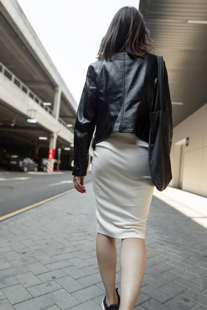 Stylowa młoda kobieta w modnych ubraniach ze skórzaną kurtką, czarną torbą na ubrania i białą sukienką spacerującą po mieście w pobliżu centrum handlowego, widok z tyłu. Moda, styl i styl życia - Zdjęcie, obraz