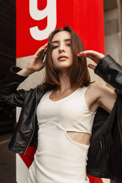 Modaya uygun kısa saçlı, siyah deri ceketli ve beyaz elbiseli güzel bir kadın model kırmızı beton duvarın yanında sokakta poz veriyor. Moda, tarz ve güzellik. - Fotoğraf, Görsel