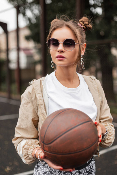 Beautiful young hipster girl with vintage sunglasses, wearing fashionable retro sportswear with hoodie and white T-shirt holding a basketball on the street. Fashion sports, style and beauty - 写真・画像