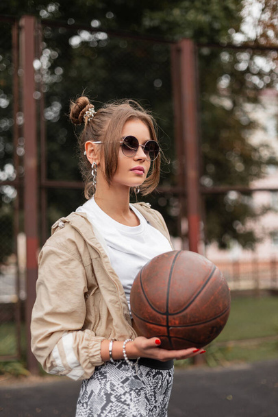 Eski moda güneş gözlüklü, modaya uygun kıyafetler giyen, rüzgarlık, tişört ve taytlı, modaya uygun güzel bir kadın basketbol sahasında topla oynuyor. - Fotoğraf, Görsel