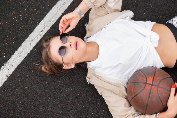 Sexy beautiful woman hipster with a ball in fashionable sportswear with a windbreaker and T-shirt, wearing sunglasses, lying and resting on the ground, top view - Photo, Image