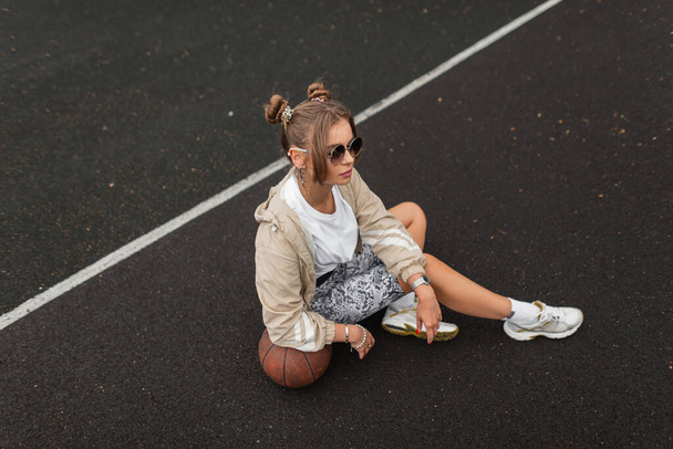 Modna, piękna, sportowa hipsterka w okularach przeciwsłonecznych w modnej wiatrówce, legginsach i koszulce z białymi trampkami siedzi i odpoczywa na asfalcie z piłką - Zdjęcie, obraz