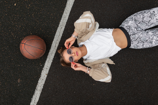 Όμορφη hipster γυναίκα στη μόδα αθλητικά ρούχα φορώντας γυαλιά ηλίου, ξαπλωμένη και χαλαρώνοντας στην άσφαλτο του γηπέδου μπάσκετ, θέα από ψηλά - Φωτογραφία, εικόνα