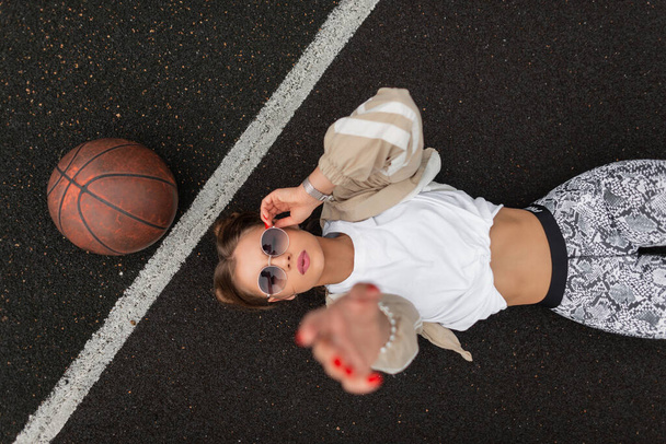 Beautiful young woman model in fashion sportswear is lying and rest on the pavement, fixing her stylish sunglasses and reaching for the camera, top view - 写真・画像