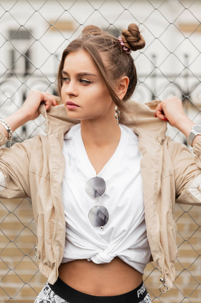 Stylová krásná mladá dívka v módní pouliční sportovní oblečení s větrovkou a bílé tričko stojí v blízkosti kovové mřížky na ulici - Fotografie, Obrázek