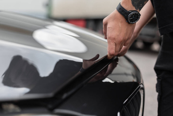 Mann öffnet Motorhaube eines schwarzen Autos und überprüft Motor und Öle. Menschenhände mit Armbanduhr öffnen die Motorhaube, Nahaufnahme - Foto, Bild