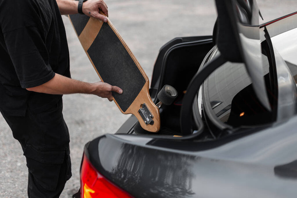 Ένας χίπστερ με μαύρα ρούχα βάζει μια σανίδα στο πορτ-μπαγκάζ του αυτοκινήτου. Ενεργά Σαββατοκύριακα και ξεκούραση - Φωτογραφία, εικόνα