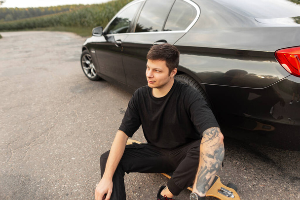 Fashionable hipster man in black T-shirt with tattoo on arm sits by car on the road outside of city - Фото, зображення