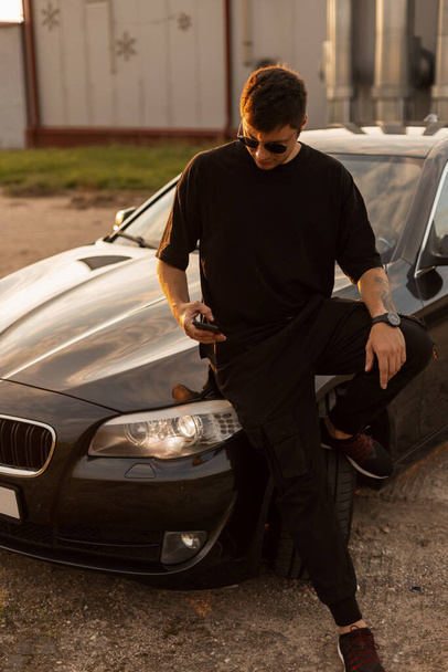 Modieuze knappe zakenman hippe vent met een coole zonnebril in zwart T-shirt en jeans in de buurt van auto zitten en kijken naar een smartphone bij zonsondergang - Foto, afbeelding