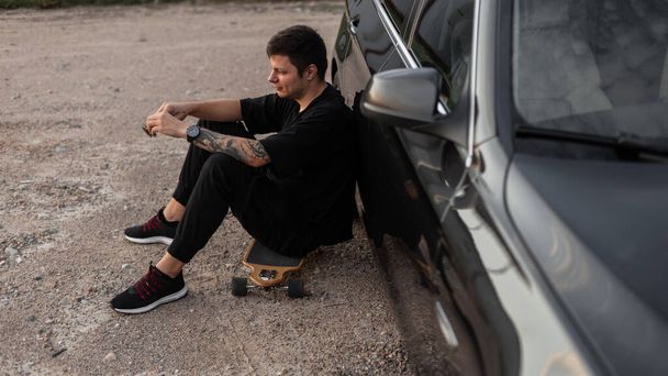 Hipster handsome man model with black clothes and sneakers sits and rest on longboard near a black car. Summer weekend - Фото, изображение