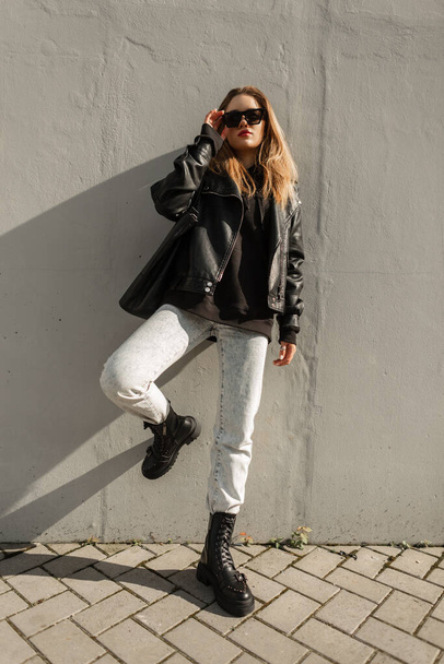 Cool fashion young beautiful woman with trendy clothes with leather jacket, pullover, jeans and shoes with bag stands and poses near a gray wall on the street. Spring, fashion and style - Fotoğraf, Görsel