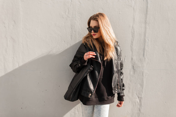 Retrato urbano de moda de mujer hermosa hipster en chaqueta de cuero de moda, sudadera con capucha y bolsa negra se encuentra cerca de una pared gris en la calle. Traje de primavera femenina, moda y belleza - Foto, Imagen