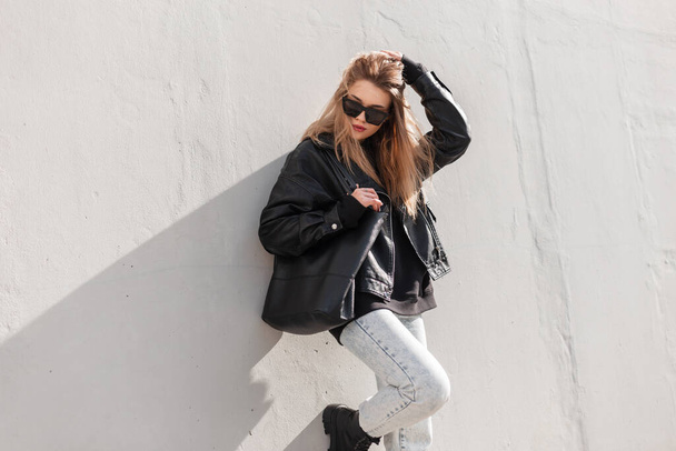 Κομψό όμορφο κορίτσι μοντέλο hipster με γυαλιά ηλίου σε μοντέρνα μαύρα ρούχα με δερμάτινο μπουφάν, κουκούλα, τζιν και μπότες με μαύρο κομψό δερμάτινη τσάντα στέκεται και θέτει κοντά σε ένα γκρι τοίχο. Αστική - Φωτογραφία, εικόνα