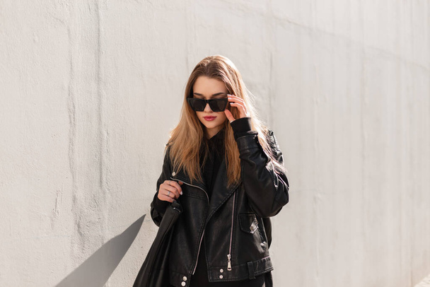 Deri ceketli, kapüşonlu ve çantalı moda güzeli hippi kız şık güneş gözlüğü takıyor ve gri bir duvarın yanında yürüyor. - Fotoğraf, Görsel