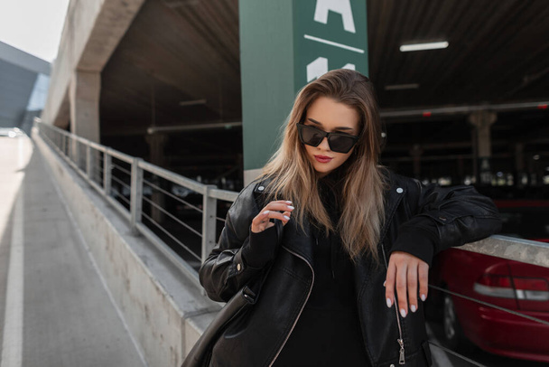 Fashionable young beautiful woman model hipster  in leather black rock jacket and black hoodie with handbag walks on the city. Urban female style look clothes - Foto, Bild