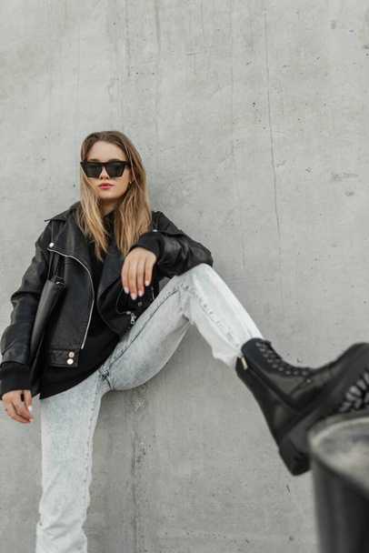 Stylish beautiful young hipster woman with sunglasses in a fashion casual outfit with a leather jacket, hoodie, jeans and boots with a bag posing on the street near a gray wall - Фото, зображення