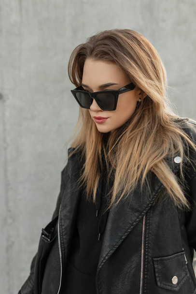 Moda modası geçmiş siyah deri ceketli ve siyah kapüşonlu şık güneş gözlüklü, saç stili ve şık bayan model hipster gri bir duvarın yanında duruyor. - Fotoğraf, Görsel