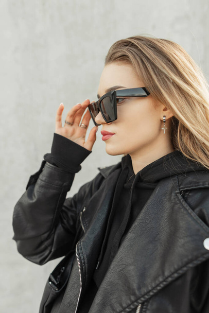 Divatos nő hipster divatos bőr fekete kabát fekete elegáns kapucnis tesz egy hűvös napszemüveg szürke beton háttér. Városi női rock stílusú ruhák - Fotó, kép