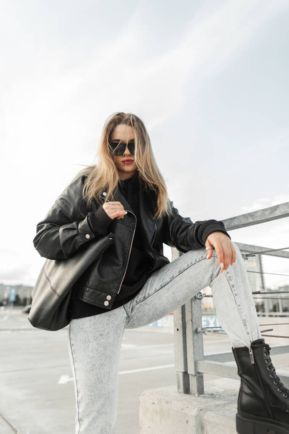 Cool fashionable young woman model hipster in fashion spring outfit with leather rock jacket, hoodie, black bag, blue jeans and leather shoes boots poses in the city. Urban female style look clothes - 写真・画像