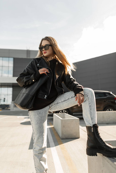 beautiful fashionable girl in black rock style with a black leather jacket, jeans, sunglasses and stylish boots with a bag poses in the city. Women's urban clothing style - Photo, image