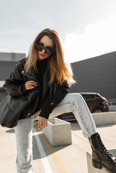 Μοντέρνο cool hipster γυναικείο μοντέλο με μοντέρνα γυαλιά ηλίου στο δερμάτινο μπουφάν μόδας, μπλε τζιν και παπούτσια με δερμάτινη τσάντα θέτει στην πόλη - Φωτογραφία, εικόνα