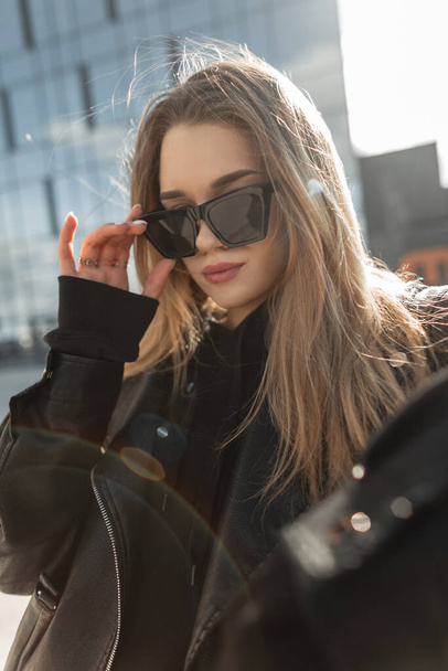 Модная красивая городская девушка-модель в модном наряде с курткой и капюшоном носит солнцезащитные очки по городу и гуляет - Фото, изображение