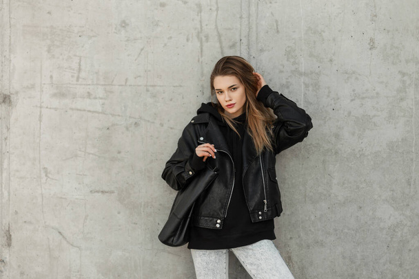 Fashion young beauty woman hipster model with rock leather jacket, hoodie and fashion leather bag stands near a concrete gray wall. Female urban style, fashion and beauty - Photo, Image