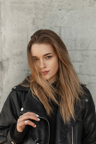 Fashionable urban portrait of young beautiful woman with hairstyle in black fashion leather rock jacket with bag stands near a gray concrete wall on the street - Fotoğraf, Görsel