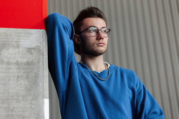 handsome hipster man in a blue sweatshirt and glasses stands near a concrete pole on the street - Foto, imagen