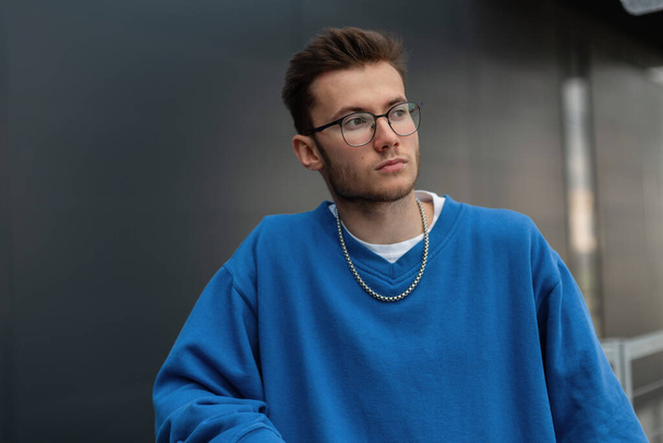 Μοντέρνος νεαρός όμορφος hipster τύπος με γυαλιά σε ένα κομψό μπλε φούτερ στην πόλη - Φωτογραφία, εικόνα