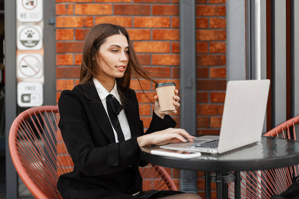 Επαγγελματική όμορφη ευτυχισμένη γυναίκα διευθυντής με μόδα μαύρο κοστούμι εργασίας σε ένα φορητό υπολογιστή και πίνοντας καφέ σε ένα καφέ κοντά σε ένα τοίχο τούβλο στην πόλη. Επιτυχημένη επιχειρηματίας κορίτσι - Φωτογραφία, εικόνα