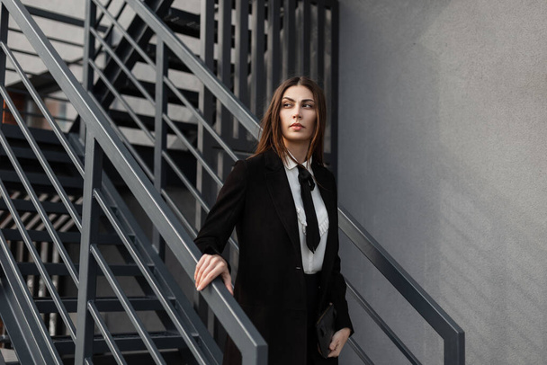 Bella donna d'affari professionale in un vestito nero fantasia con una camicia, cravatta e cappotto cammina giù per le scale di metallo fuori in un edificio per uffici - Foto, immagini