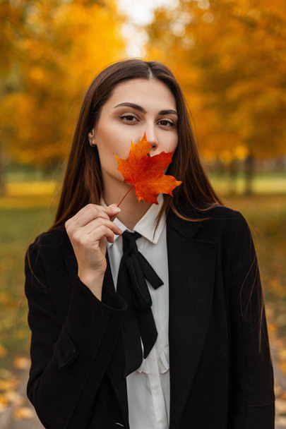 Şık iş kıyafetleri içinde güzel bir esmer kadın renkli sonbahar parkında yürüyor ve yüzünü kırmızı-turuncu bir akçaağaç yaprağıyla kaplıyor. - Fotoğraf, Görsel