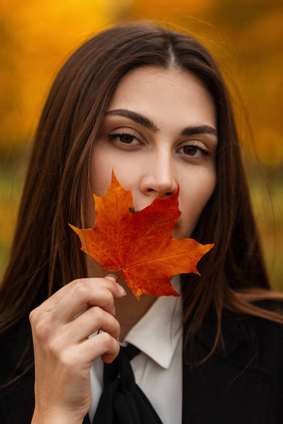 Stylisch schöne junge Frau in modischen schwarzen Kleidern bedeckt ihre Lippen mit einem herbstlichen Ahornblatt und spaziert durch den goldenen Herbstpark - Foto, Bild