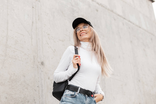Divertido hermosa mujer joven con una linda sonrisa con una ropa casual de moda con una gorra, gafas, suéter blanco y una mochila está caminando en la calle cerca de una pared gris de hormigón. - Foto, imagen