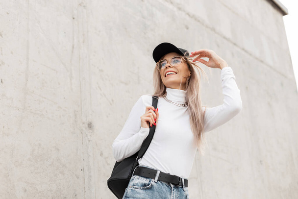 Feliz hermosa chica divertida con una sonrisa en ropa casual de moda con una gorra negra y una mochila está caminando por la calle cerca de una pared de hormigón gris - Foto, Imagen