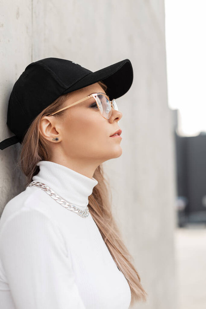 Fashionable urban portrait of a beautiful young woman with glasses in a fashionable black cap and white sweater stands near a concrete gray wall in the city - Zdjęcie, obraz