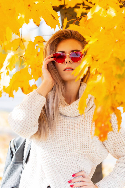 Chica joven con estilo fresco con gafas de lujo en un suéter blanco vintage con mochila se encuentra en el follaje de otoño de oro brillante en el parque - Foto, Imagen