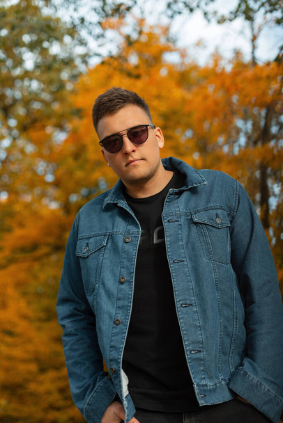Stylish handsome young man with trendy sunglasses wearing a blue denim shirt with a black tank top and jeans in an autumn park with yellow foliage - Fotoğraf, Görsel