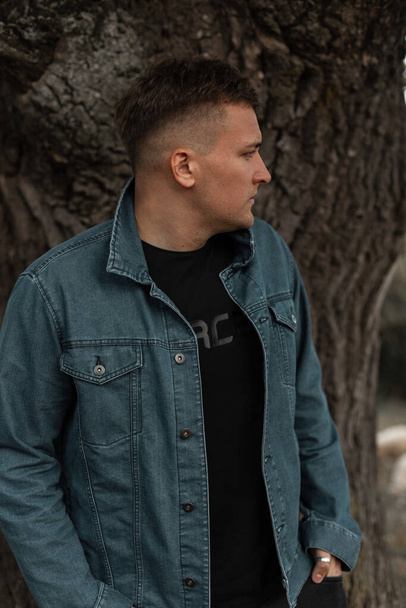 Fashionable handsome guy with his hairstyle in a stylish denim jacket in the countryside near a tree - Фото, зображення