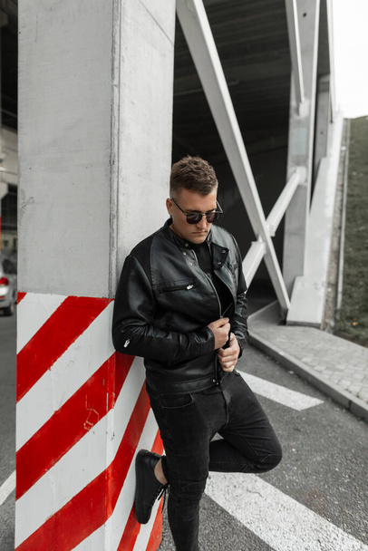 Guapo chico urbano con estilo fresco en ropa negra de moda con gafas de sol, chaqueta de cuero, camiseta y jeans se para y posa cerca de un poste de hormigón en la calle - Foto, imagen