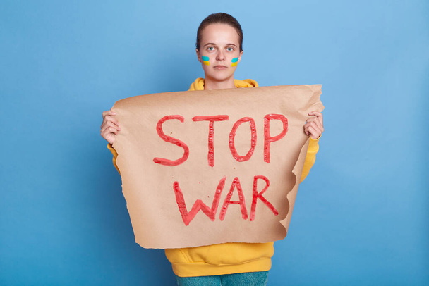 Portrait of a beautiful Caucasian young adult woman holding a sign with the inscription Stop War, showing her support for the country during war, posing isolated over blue background. - Foto, Bild