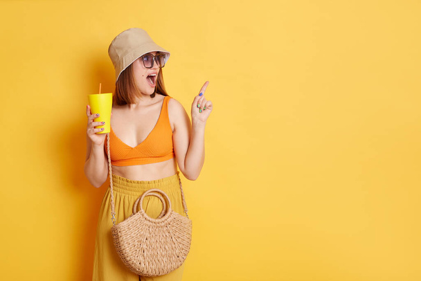 Vnitřní záběr užaslé ohromený žena v slunečních brýlích na sobě letní oblečení pózovat izolované přes žluté pozadí, stojí s čerstvým v hadns a ukazuje stranou a reklamní plochy. - Fotografie, Obrázek