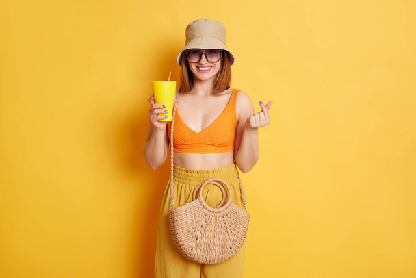 Image of lovely good looking woman in sunglasses wearing summer clothing posing isolated over yellow background, enjoying juice and showing korean love symbol. - Фото, зображення