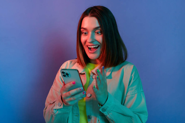 Portrait of amazed surprised Caucasian woman wearing stylish jacket posing isolated on neon light background, using cell phone, reading breaking news, looking at display with big eyes. - Фото, изображение