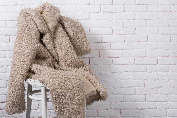 beige faux fur jas, als zittend op een stoel tegen een witte bakstenen muur, kopieer spase - Foto, afbeelding