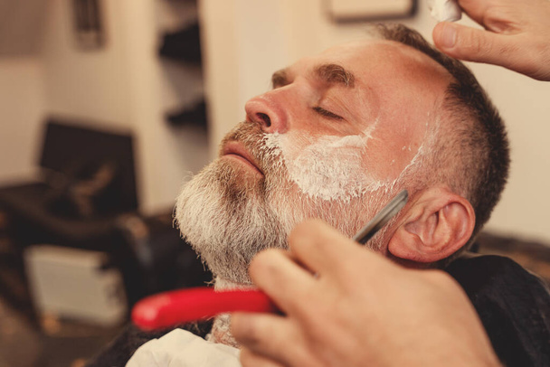 Afeitado con una navaja de afeitar en una barbería. Un viejo barbudo afeitado en una barbería. Afeitado clásico de acero inoxidable Razor borde recto. - Foto, imagen