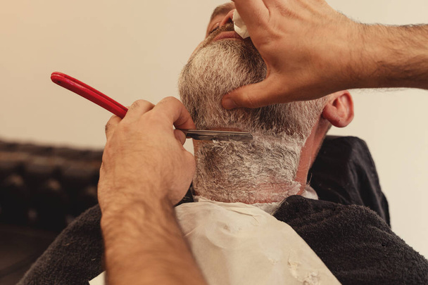 Shaving with a straight razor in a barbershop. A bearded old man being shaved in a barbershop. Classic shave by Stainless Steel Straight Edge Razor. - Fotoğraf, Görsel