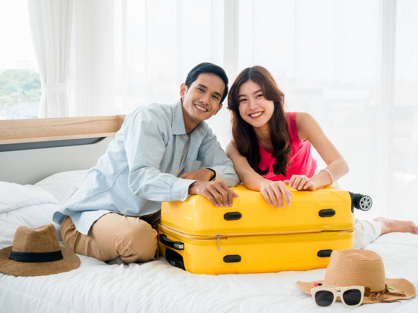 Couple travelers preparing to trip. Asian man in denim shirt and young beautiful sitting together and packing bag, yellow suitcases on bed in bedroom. Happy holiday. Colorful summer vacation. - Photo, Image