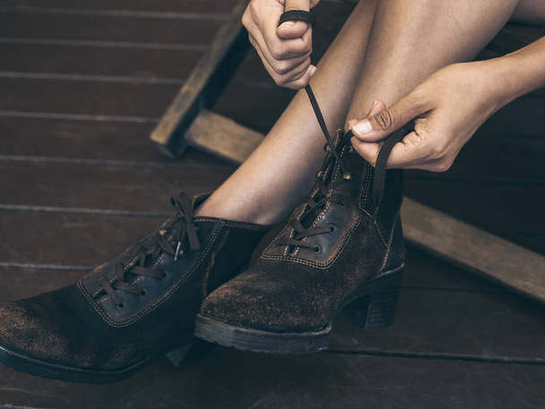 Egy közeli nő kezei, amint cipőfűzőt kötöznek a lábában lévő régi bőrcsizmára, miközben fából készült széken ülnek a deszkás teraszon.. - Fotó, kép